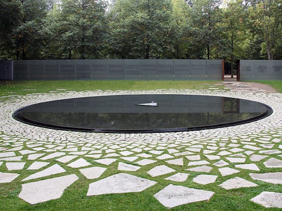 A black pool surrounded by white flagstones. In the center of the pool is a triangle with a single flower on it.