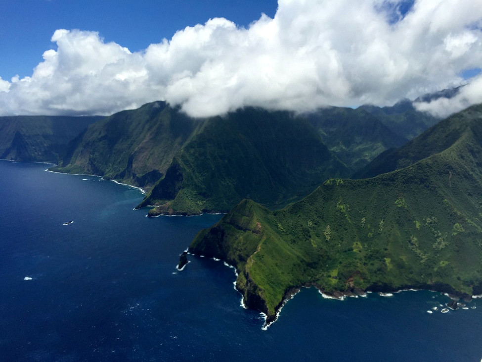 An aerial view of the north shore on Molokaʻi, Hawaiʻi, showing the coastline, green mountains, and blue ocean.