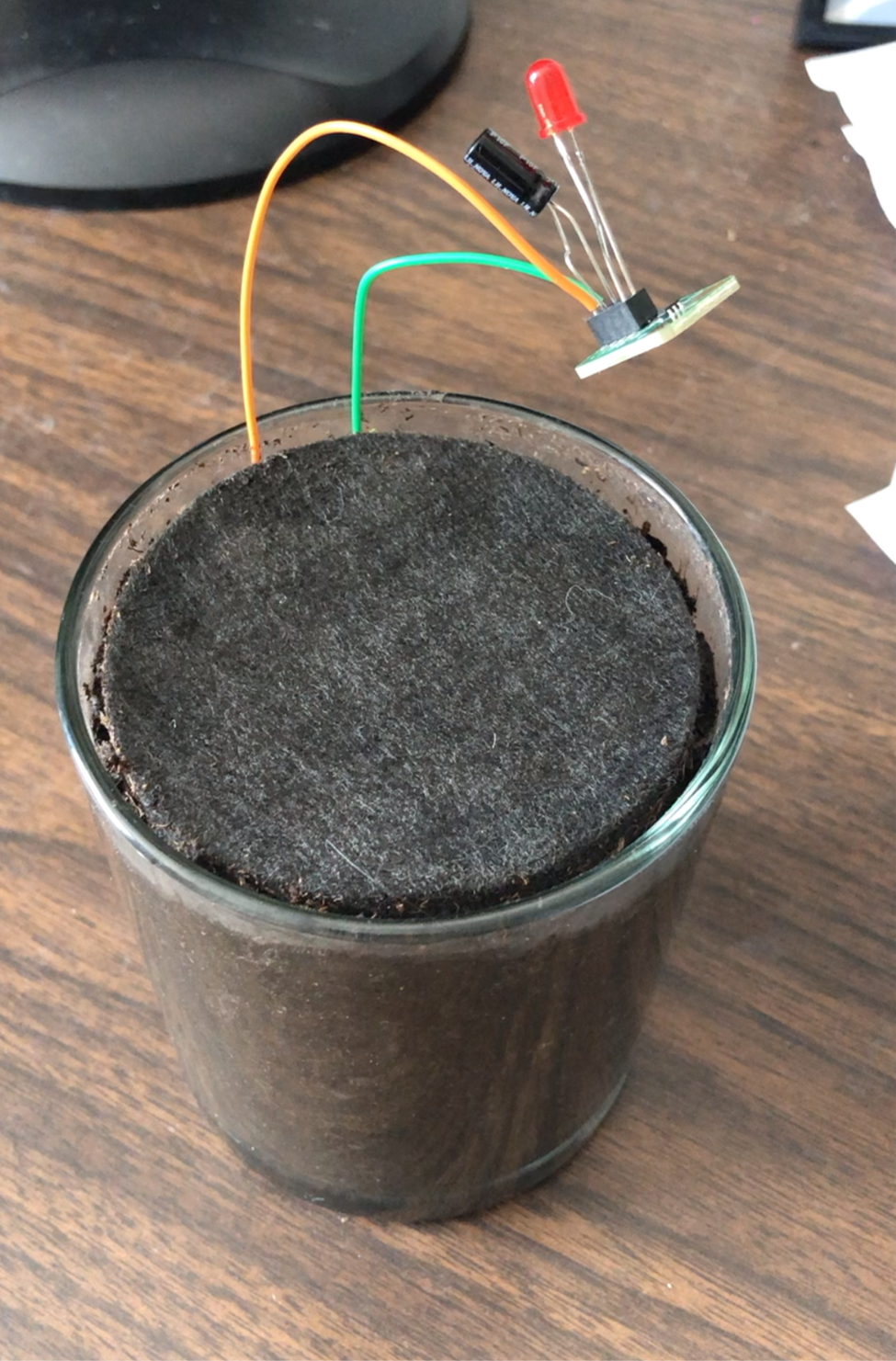 An image of soil in a small glass vase with wires and a blinker LED board coming out of it.