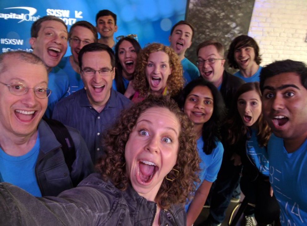 Selfie of a dozen smiling people wearing the same light blue t shirt.
