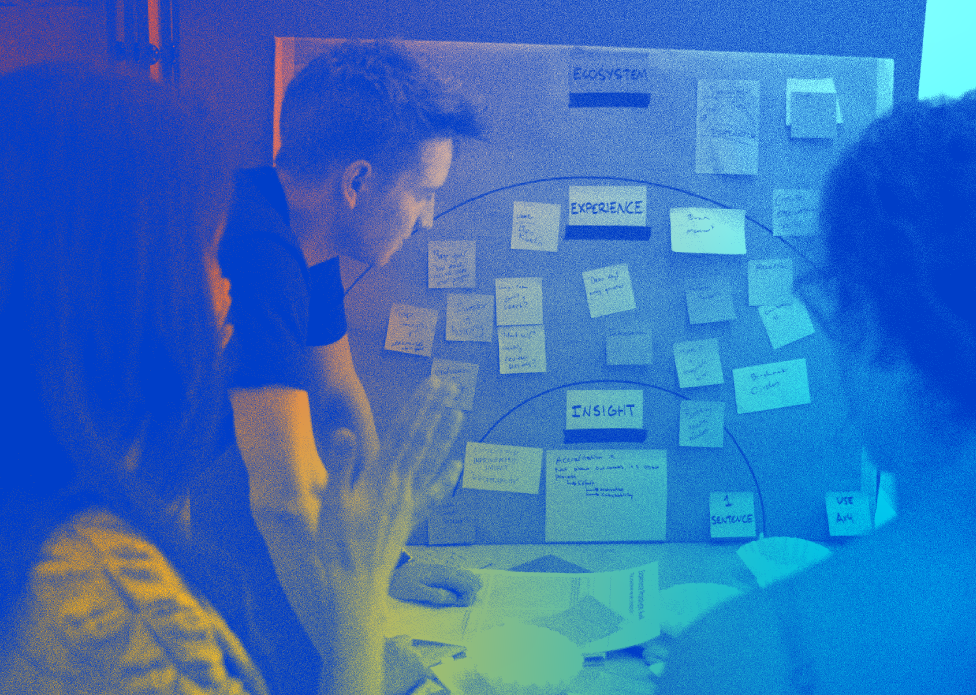 A blue and gold colored image of a group of three people standing together and working on a radial arrangement of sticky notes. The center radius is labeled “insight” the next outward ring is titled “experience” and the widest-encompassing ring reads “ecosystem.”