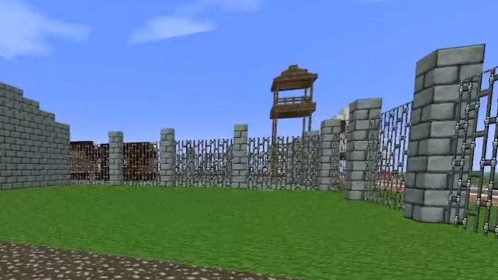 A pixelated grasssy area in Minecraft fenced in using stone pillars and a chain link fence. There is also a watch tower made of wood.