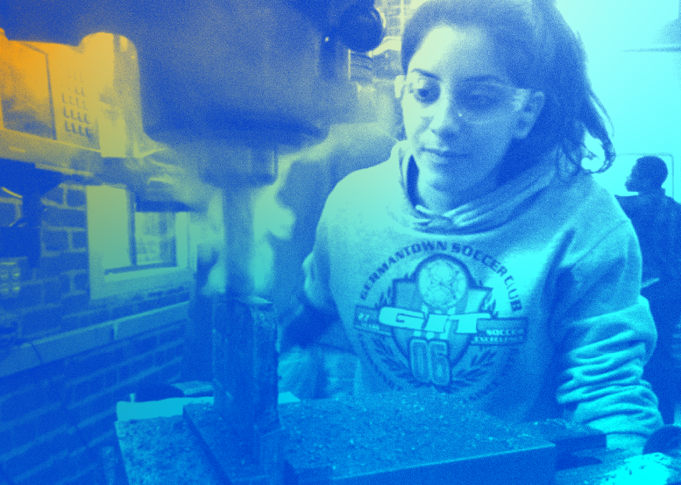 Psychedelic light blue and gold coloring of an image of young, baby-faced Tania Anaissie wearing a sports-themed sweatshirt and safety goggles. Tania is working with a piece of bronze metal on a mill machine, with a slight smile and gaze focused on the spinning machinery.