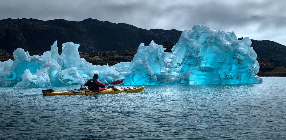 A sea kayaker paddles past a beautiful, blue-tinted iceberg