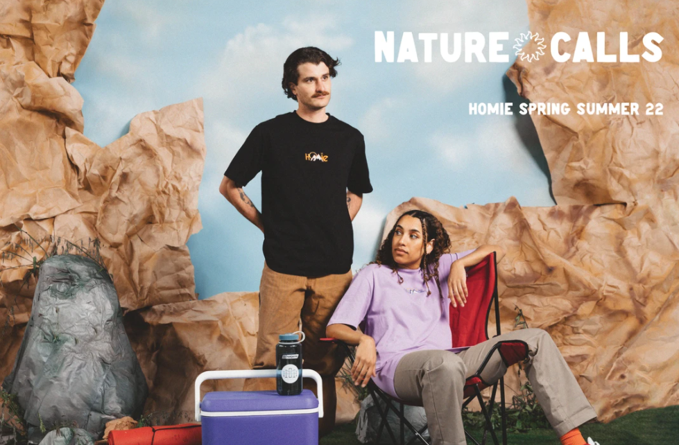 Two models are wearing Homie clothes in front of a mockup camping set. A male is standing with his hands on hips looking into the distance. He wears a black t-shirt with ‘Homie’ on the front. A female is sitting next to him on a camping chair with her legs draped over the arm of the chair. She wears a purple t-shirt with ‘Homie’ on the front.