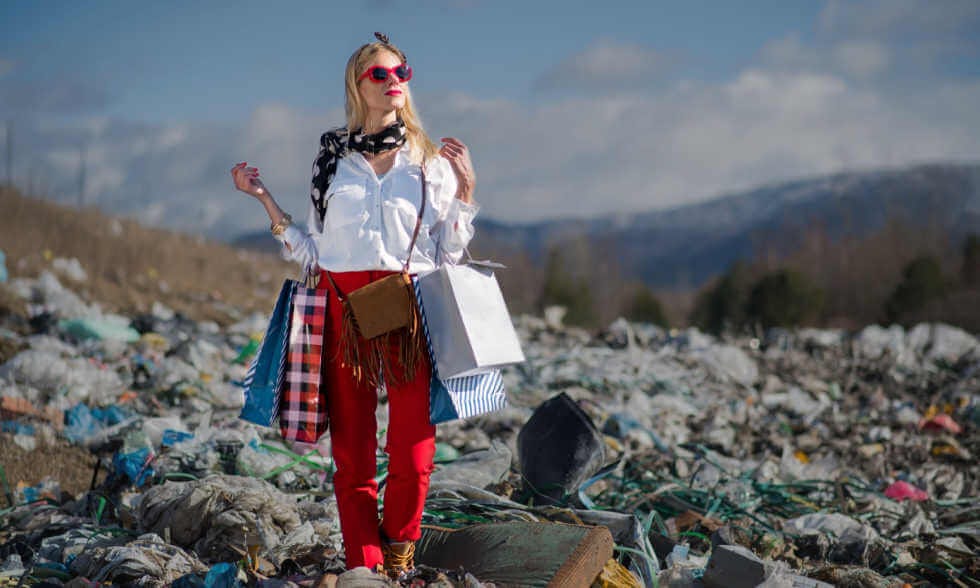 Image of a woman with shopping bags standing in a landfill of waste clothes