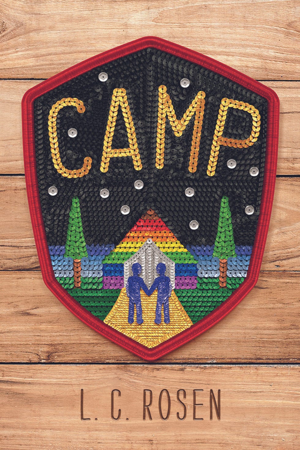 The cover of Camp by LC Rosen. The background is lightly colored wood planks, and a large sequined badge takes the foreground. Camp is spelled out in gold sequins in a starry night. On the gorund level, two blue-sequined figures hold hands in front of a gay rainbow colored building.