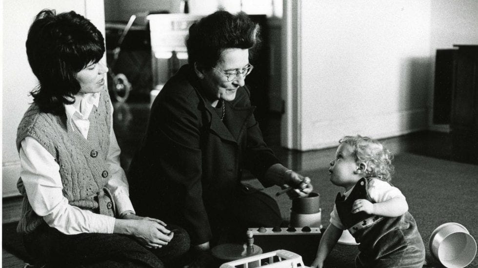 A black and white image shows Mary Ainsworth conducting her study. She sits on the floor looking towards a toddler, whose mother is sitting with them off to the side.