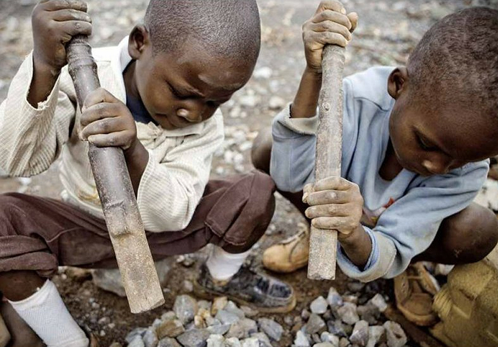 Two young boys sitting on the ground, using large hammers to break cobalt stones.