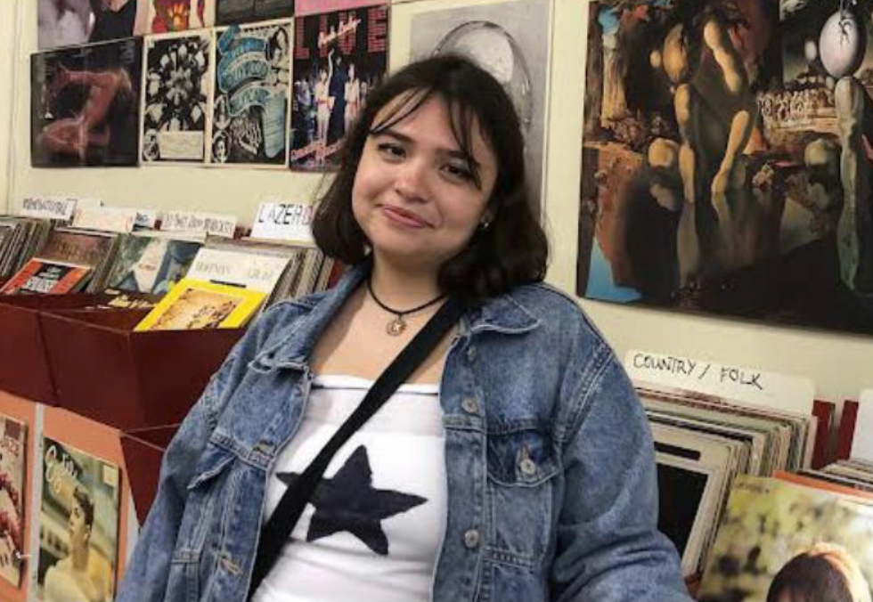 Sarah Licón posing in front of a shelf of music records. Behind her is a while wall covered with posters. She is wearing a denim jacket and a white shirt with a star at the center, a cross-body bag strap on her shoulder.