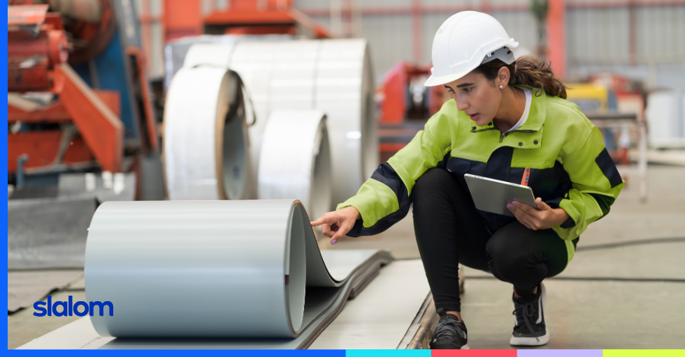 Woman in a hardhat in a warehouse crouching beside a roll of manufactured material