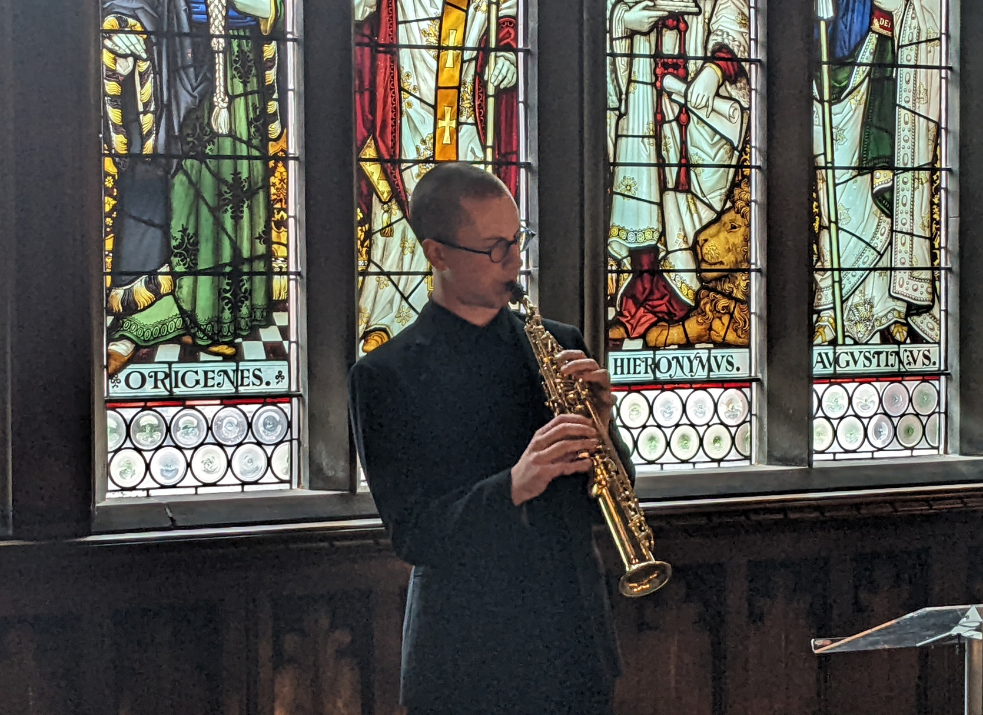 Saxophonist plays on a balcony with a fine stained glass window behind him