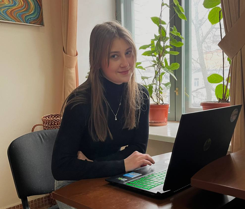 A young woman in a black sweater sits at a desk next to a window, typing on a laptop.