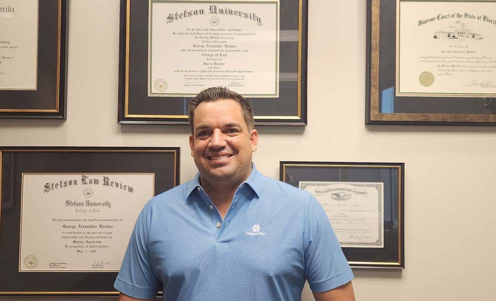 A man in a blue shirt stands proudly in front of framed certificates, showcasing his achievements and qualifications.