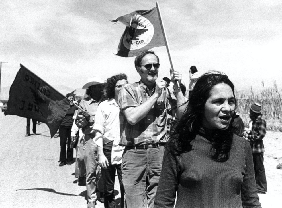 Group of people (7 or more visible) marching on a road, holding flags. Flag is from the United Farmer Workers Union.