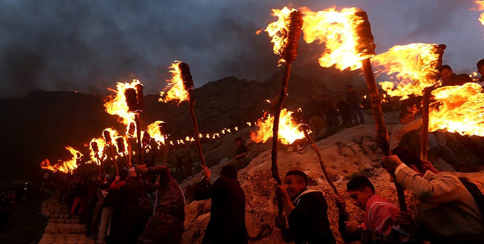 A darkened image (whether by smoke or dusk) of people in Iraqi Kurdistan waving burning sticks of fire in celebration. They are surrounded by rocks.