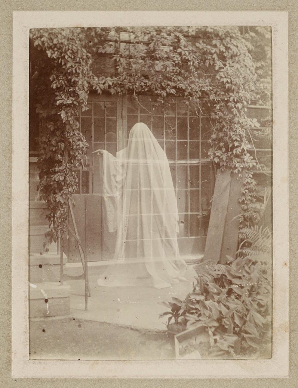 A white-translucent sheet-covered ghost, in front of a glazed and overgrown building entrance, points left at something beyond the photo’s frame.