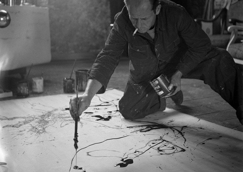 Image of Jackson Pollock painting on the floor with a brush, can of paint and cigarette.