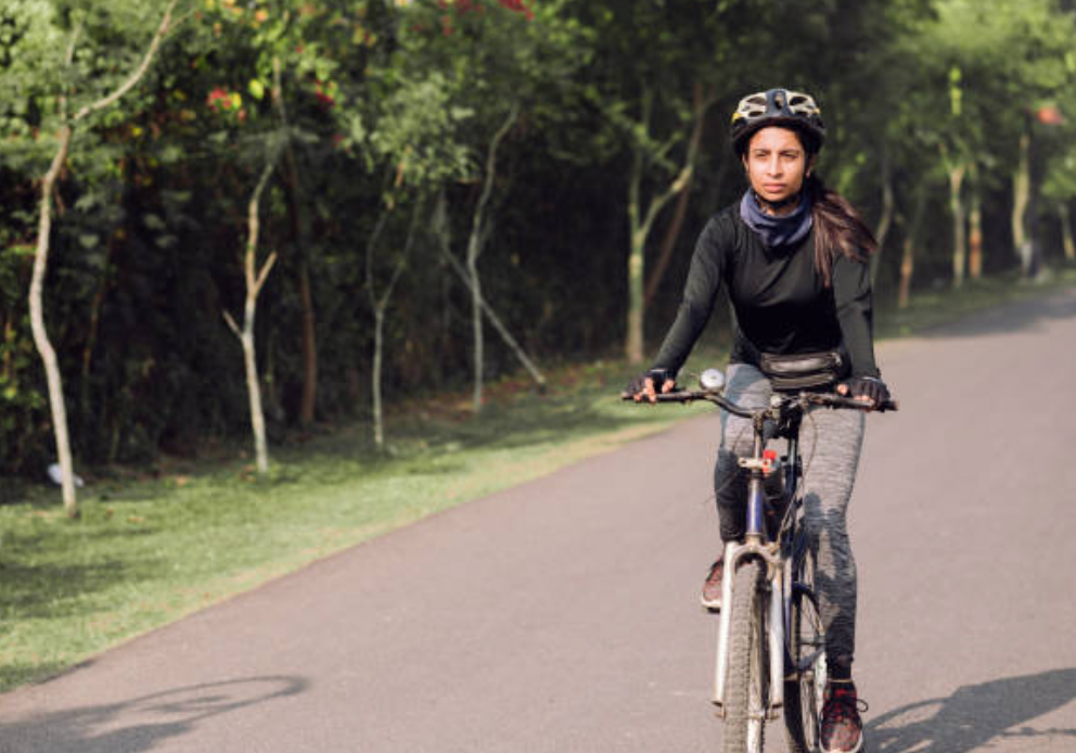 An Indian cyclist rides her bicycle on an empty street surrounded by trees on one side, during daytime.