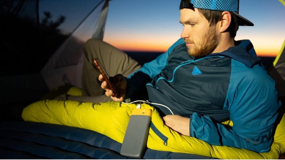 Man laying down relaxing in his tent at night while camped out somewhere. He is looking at his phone that is plugged into a portable charging device.