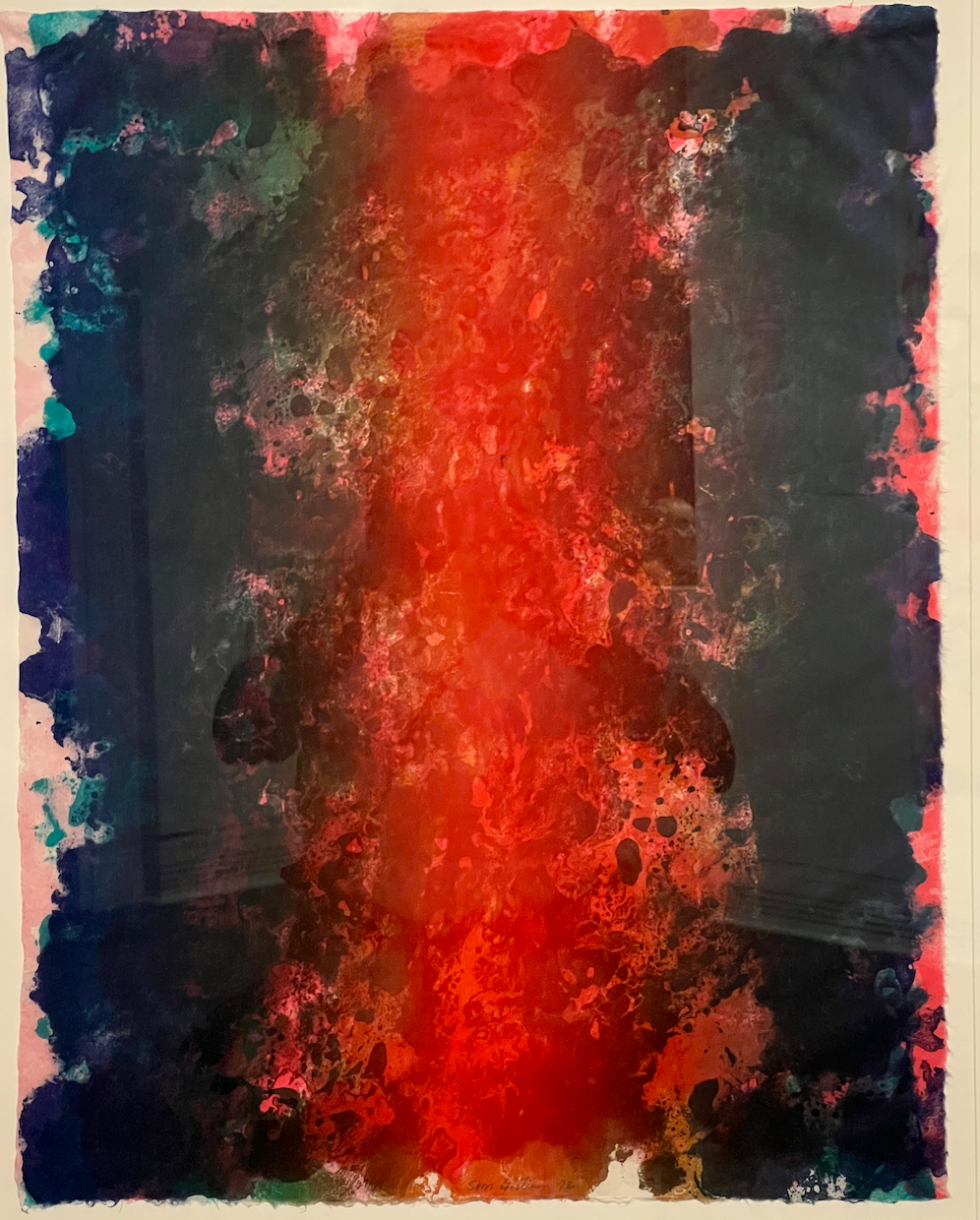 An abstract painting by Sam Gilliam. The painting has various shades of reds and blues. It hangs on a white gallery wall. Candace, who took the picture, is slighty visible in the reflection of the glass frame.