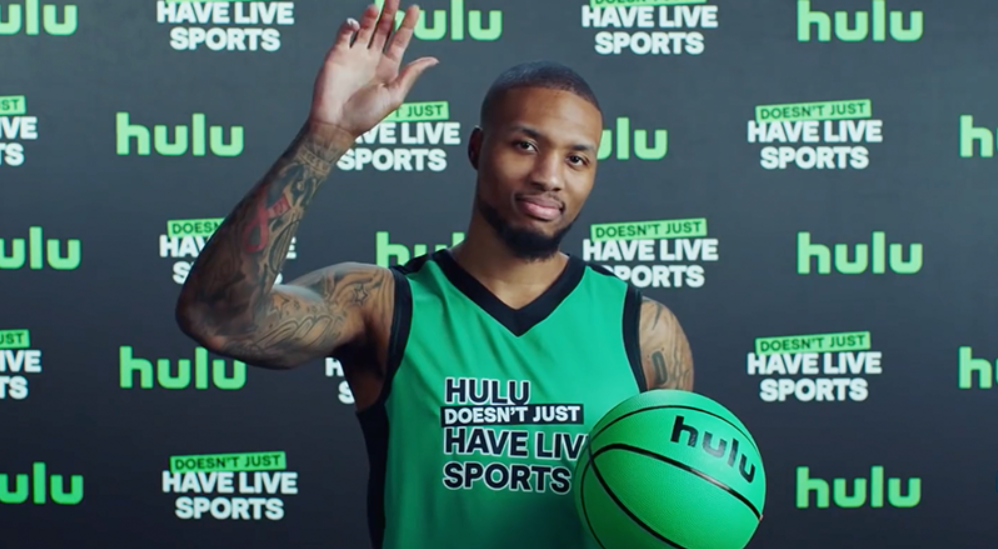 Photo of Damian Lillard wearing a “Hulu Doesn’t Have Live Sports” basketball jersey, holding a green ‘Hulu’ basketball & waving to the camera.