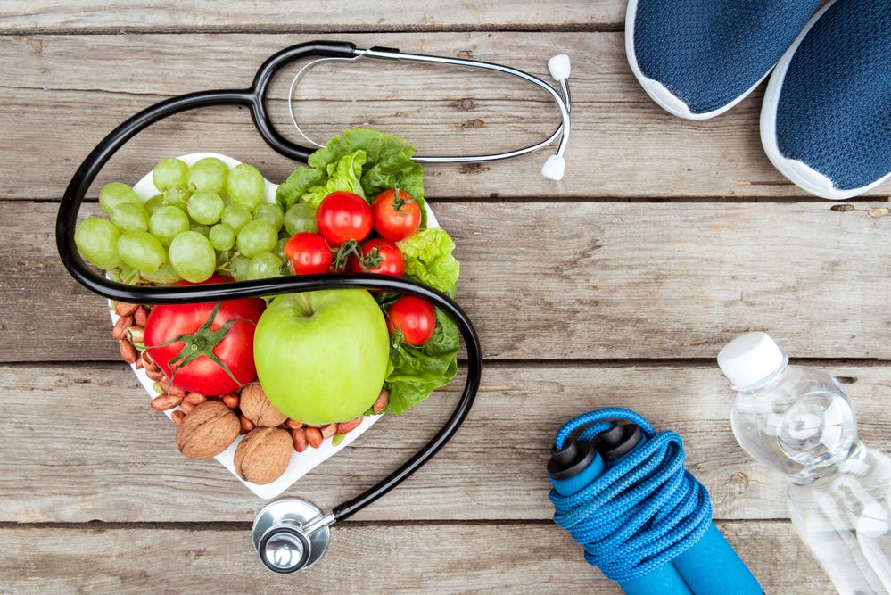 A heart shaped bowl of fruits and vegetables with a stethoscope draped across it. A pair of sneakers, a water bottle, and folded jump rope sit nearby.