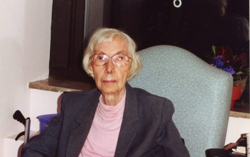 Eileen Egan as a white-haired woman, seated in a chair in her home wearing a black blazer and pink crew neck top.