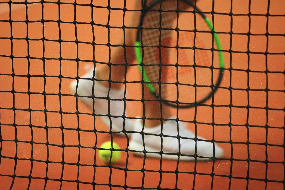 Rede de tênis em primeiro plano, e detrás dela vê-se as pernas de uma pessoa branca, que usa tênis brancos, e segura uma raquete. Ao lado, no chão, há uma bola de tênis.