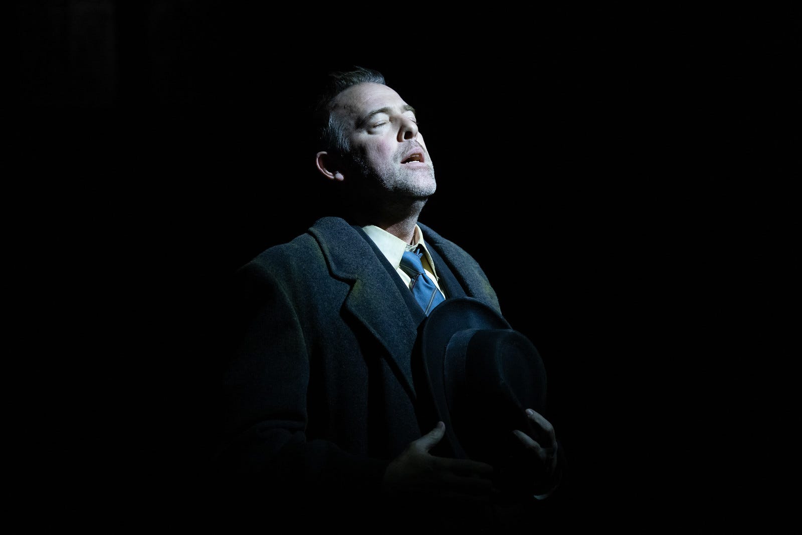 A man wearing a dark suit stands in a spotlight, eyes closed and holding his hat to his heart