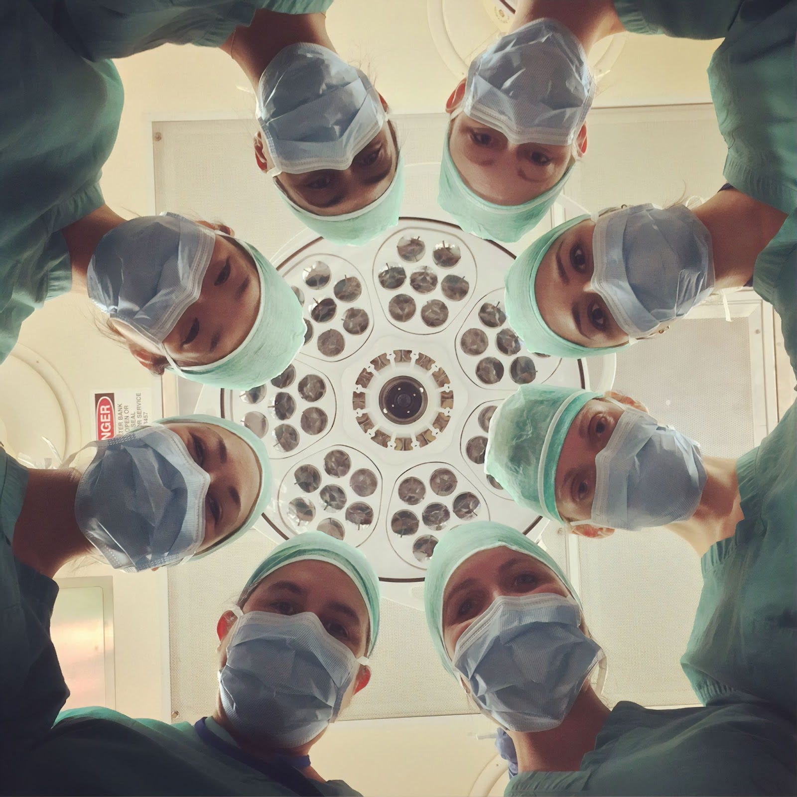 A photo of eight medical professionals wearing light green scrubs, matching caps, and blue surgical masks. Most of their faces are hidden under the masks, with only their eyes visible. They are standing in a circle and looking down at the camera, which takes the position of a patient lying on a hospital bed. Above the doctor’s head is a set of lights and the ceiling, both of which indicate that the room is a surgical theater.