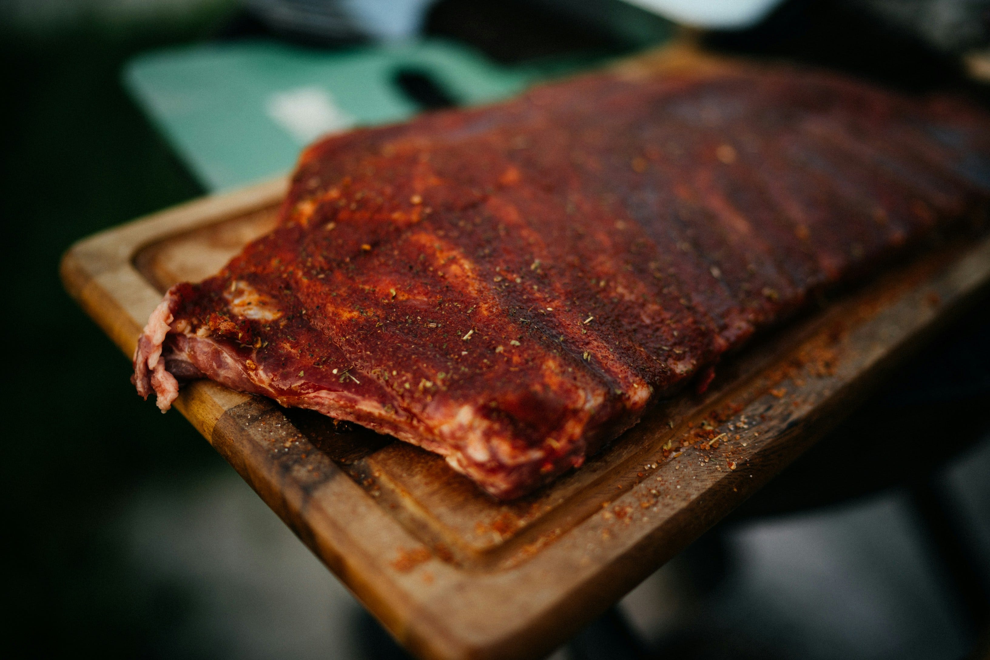 A close-up color photo showing a full rack of pork ribs sitting on a cutting board, dark red in color from rubbed on spices and sauce.