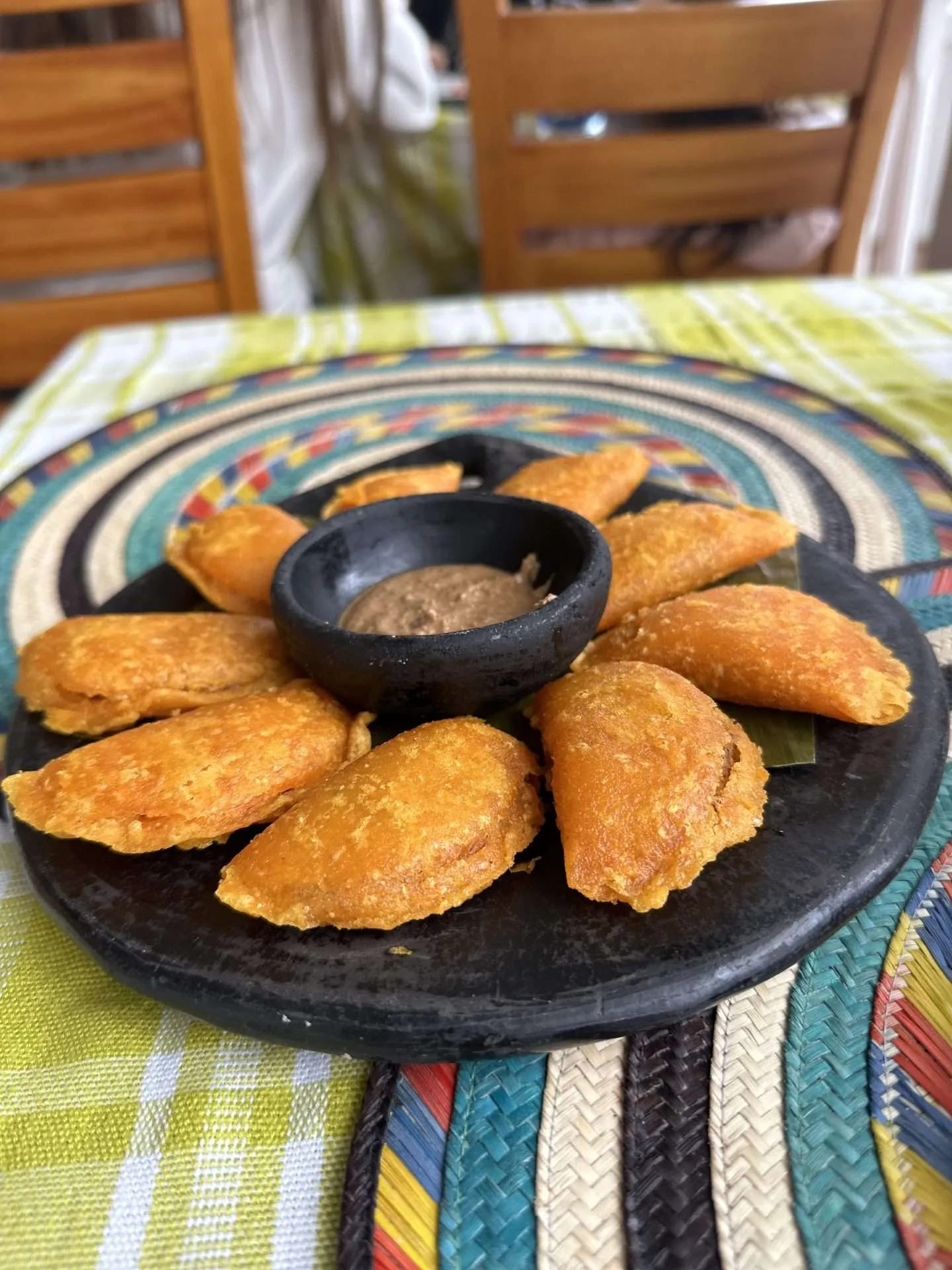 A plate of golden-brown empanadas arranged around a central dipping sauce, placed on a decorative woven mat and tablecloth—a perfect meal after pivoting during full-time travel. Wooden chairs are visible in the background.