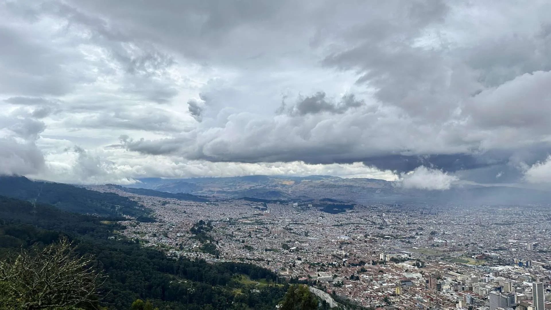 A panoramic view of a city sprawled over a flat valley with dense urban buildings, surrounded by green hills under a cloudy sky; perfect for those pivoting during long-term travel to experience both nature and urban environments.