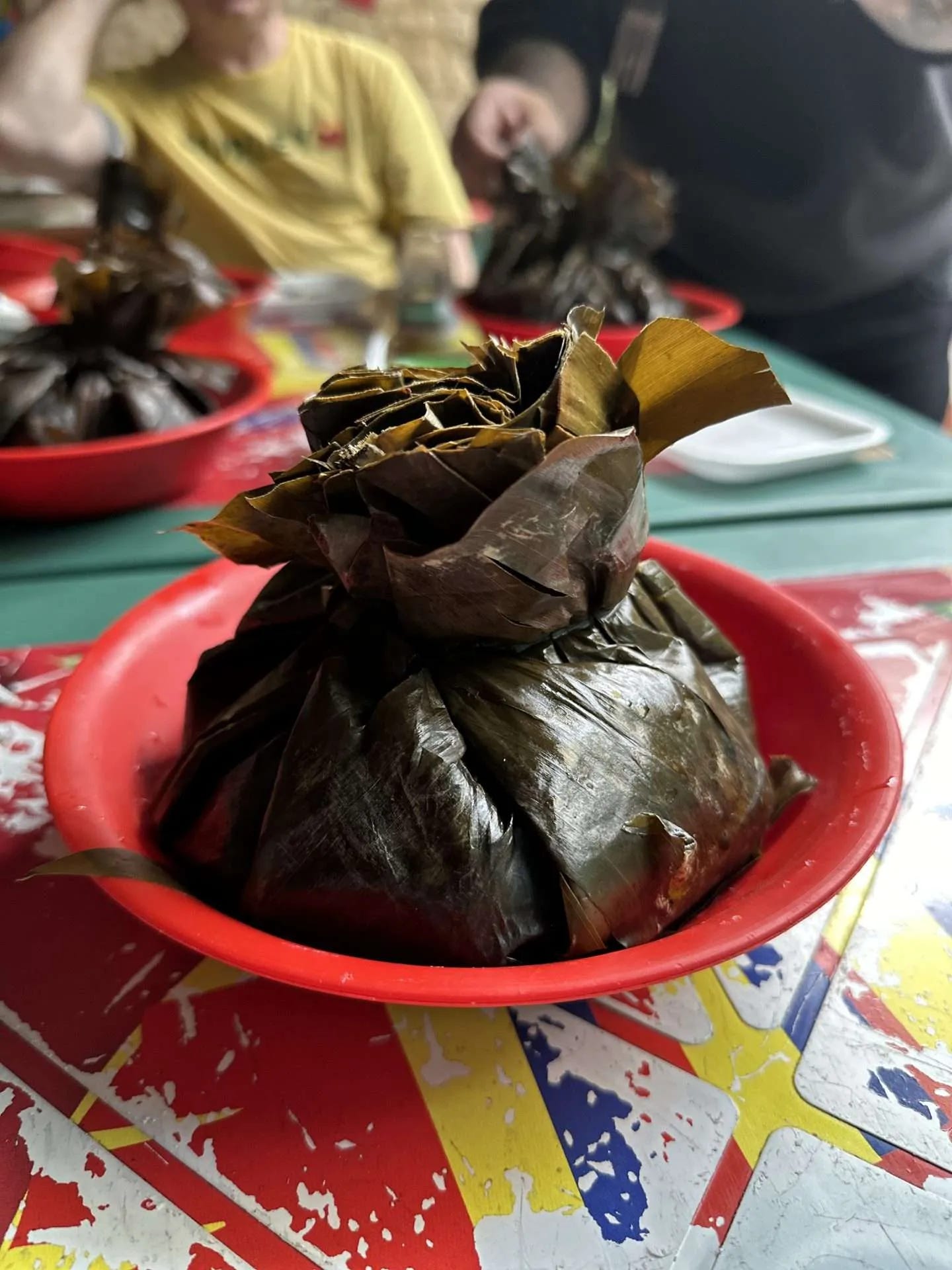 A food item wrapped in banana leaves is placed in a red bowl on a table alongside other similar dishes, exemplifying the art of making changes on the go. Two people are visible in the background, perhaps pivoting during full time travel to savor new culinary delights.