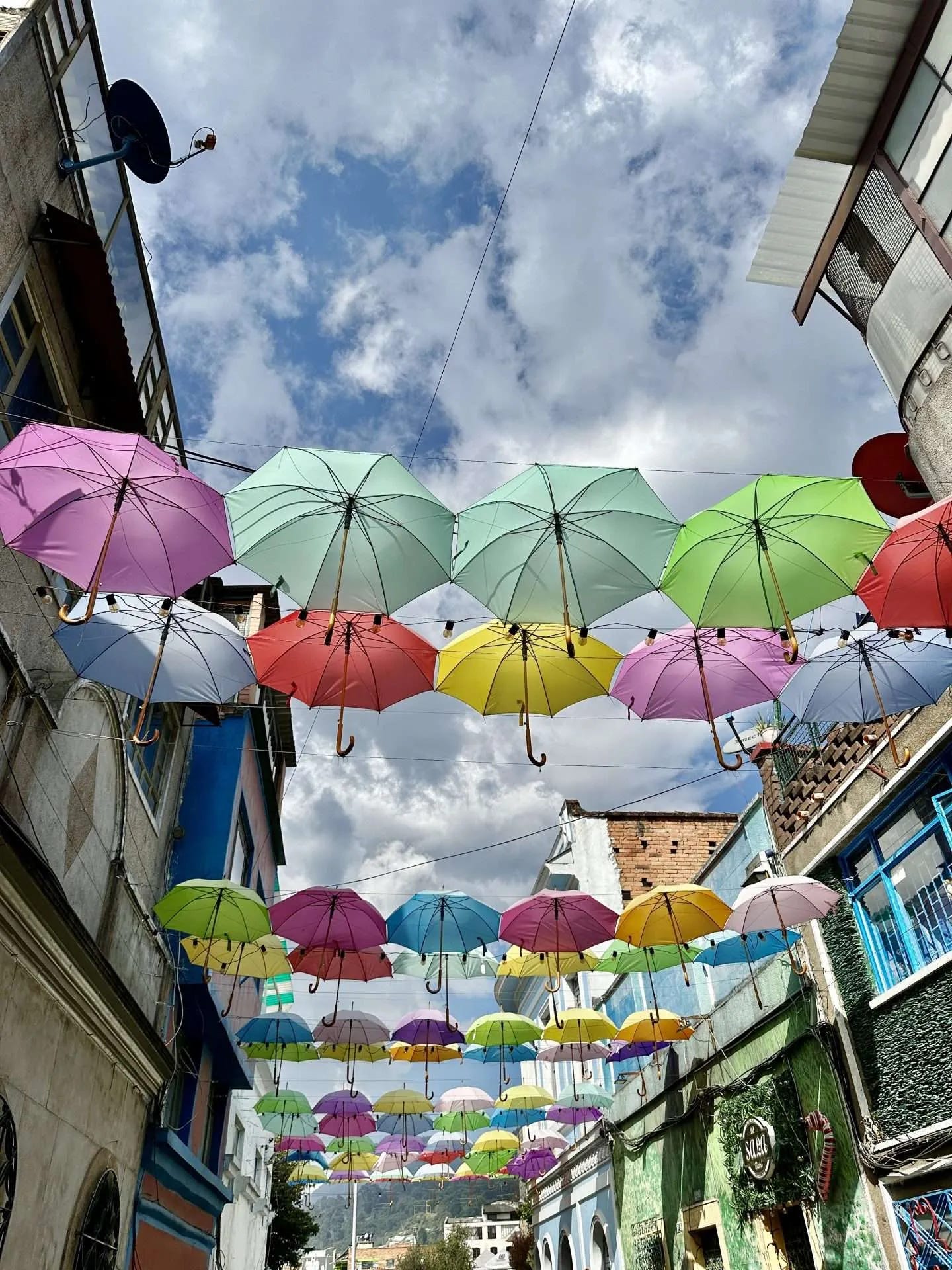 Colorful umbrellas hang suspended over a narrow street between buildings, with a partly cloudy sky in the background, capturing the whimsical essence of full-time travel.