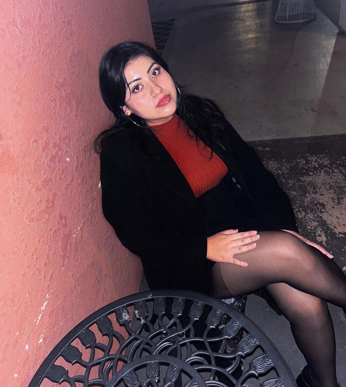 Shelby Silva is sitting outside and leans against a pink wall. She has her legs crossed and she’s wearing a black blazer over a red knit sweater. She wears a black skirt and sheer leggings. There is an iron table next to her.