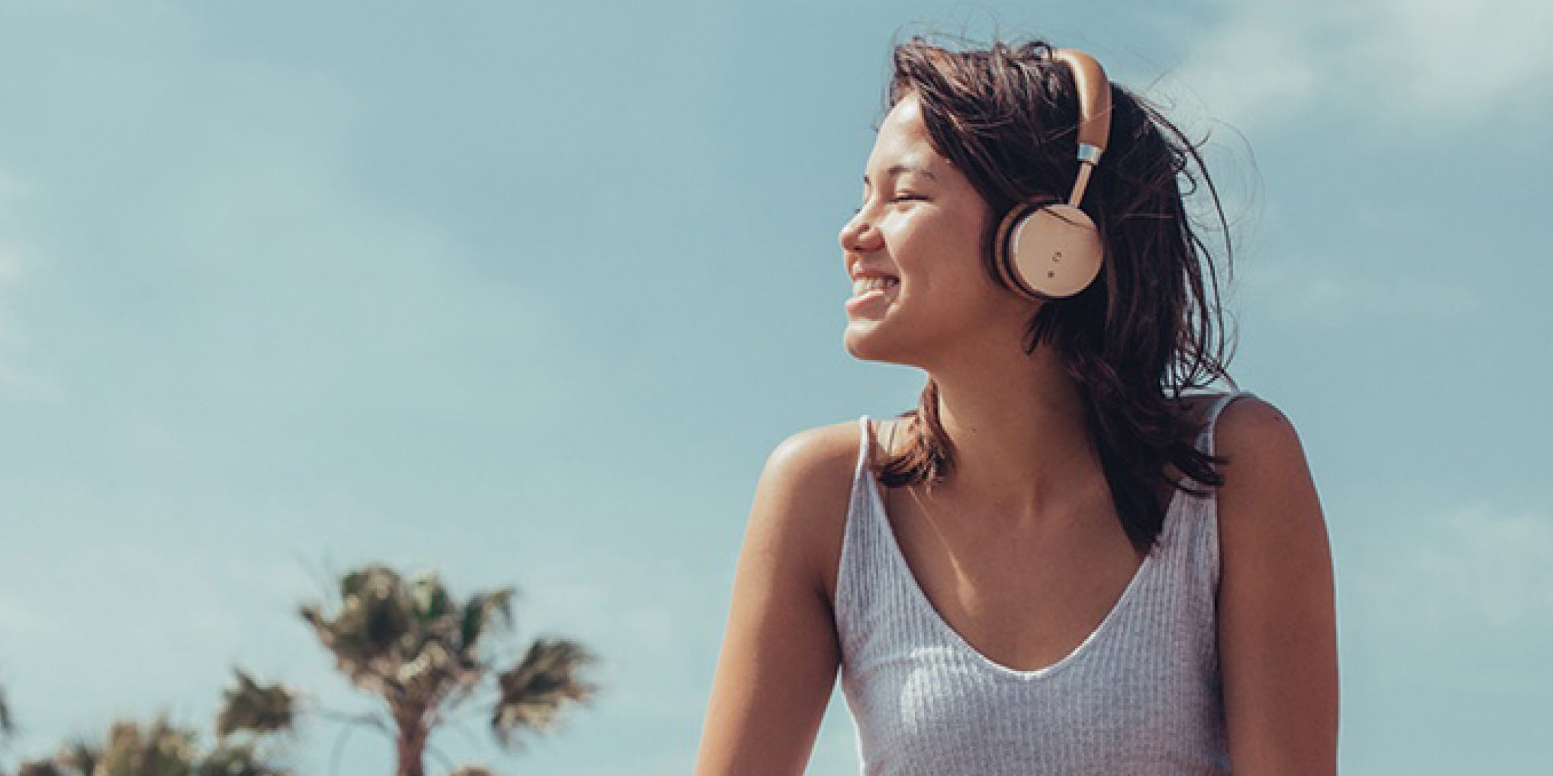 A woman wearing a tank top is with her eyes closed and is wearing headphones while smiling and facing the sunlight.