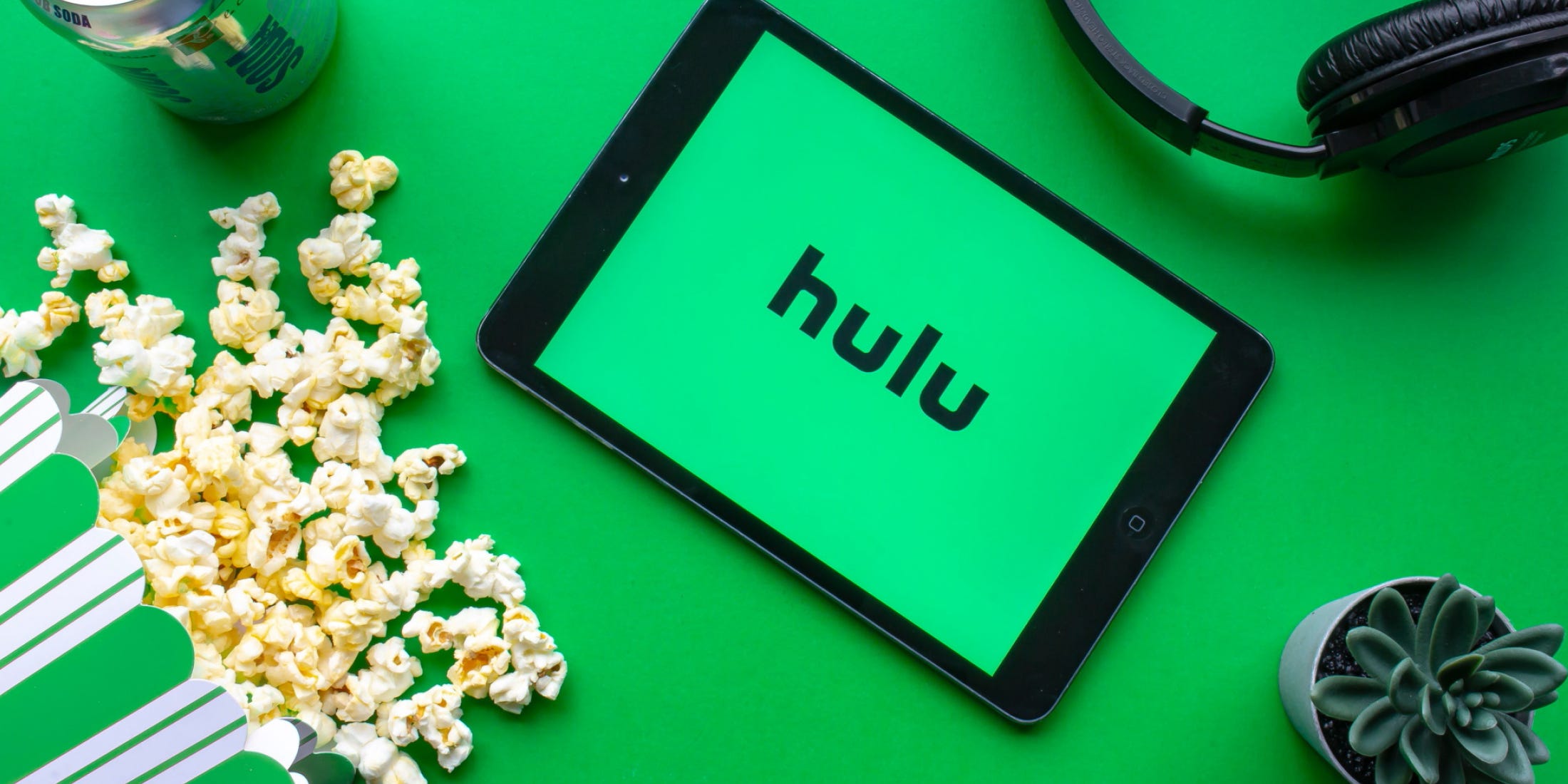 A tablet with a green background and the word “hulu” lays on a green table. It is surrounded by spilled popcorn, a succulent plant, a green soda can, and black headphones.