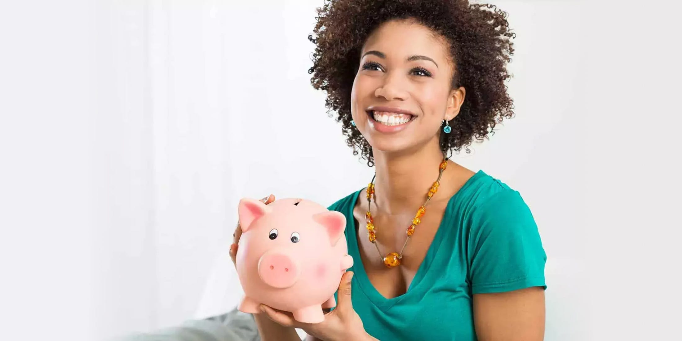 A woman with curly hair and a teal blouse is looking up and smiling while holding a classic, pink piggy bank.