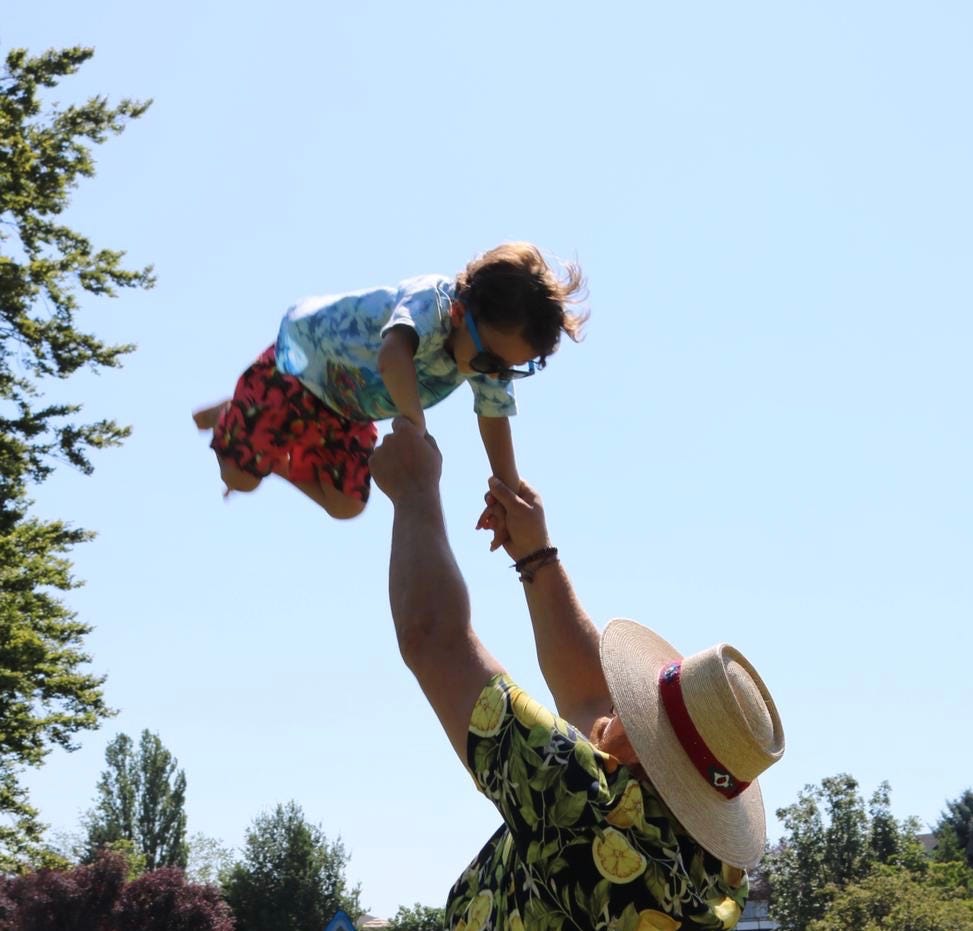 A 3 year old boy with dangling sunglasses is suspended high above his uncle holding him up with outstretched arms.